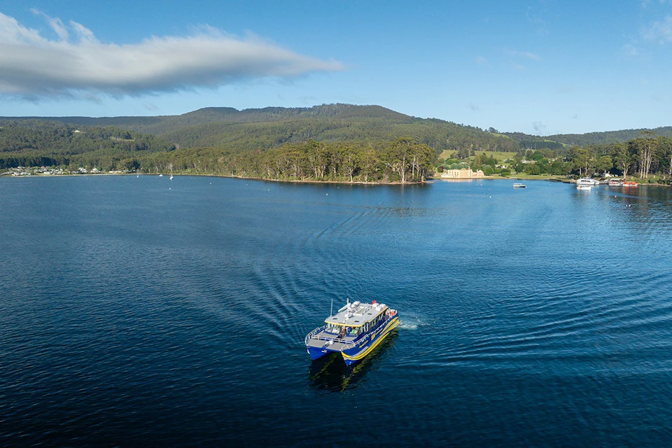A blue and yellow boat sails on a calm lake, with a tree-lined shore and distant hills under a blue sky.