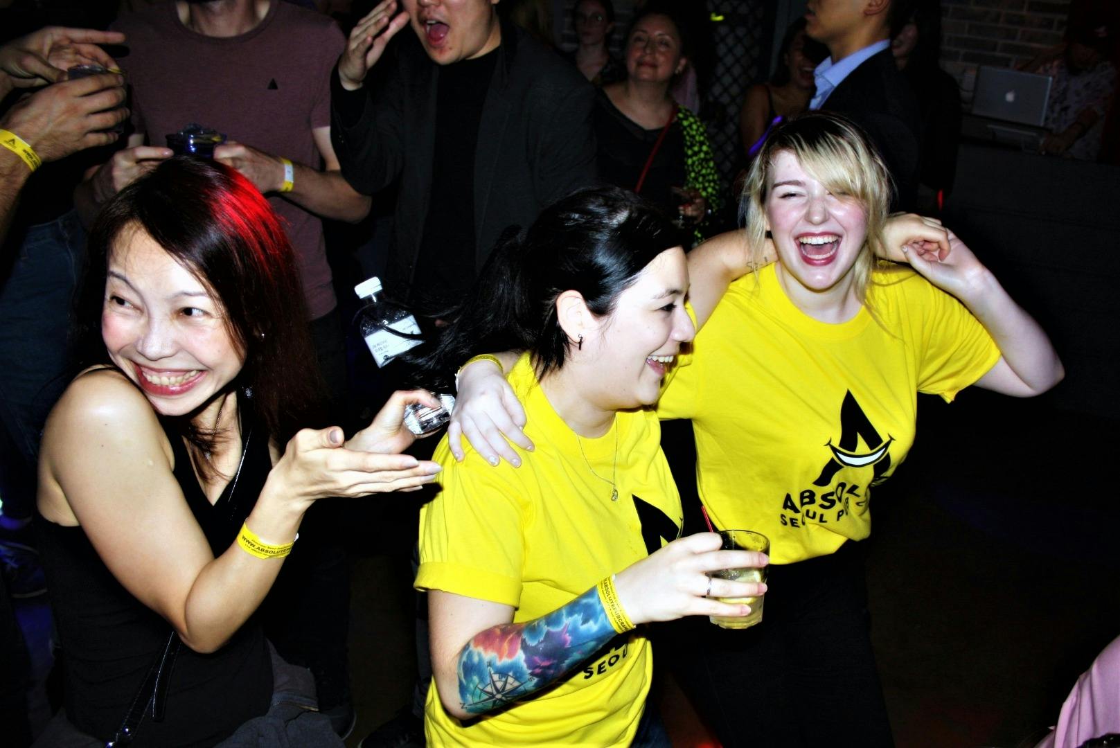 A lively group of people, including two women in yellow shirts, smiling and laughing in a crowded indoor setting.