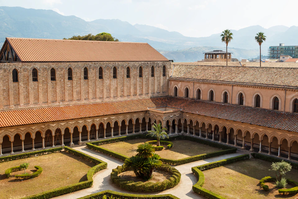 A courtyard with trimmed hedges and palm trees, surrounded by arched cloisters and buildings with terracotta roofs and mountains in the background.