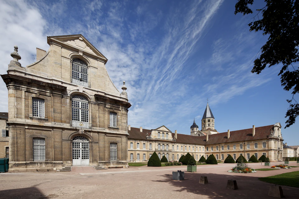 A historic building with arched windows and a courtyard featuring manicured shrubs, under a partly cloudy blue sky.