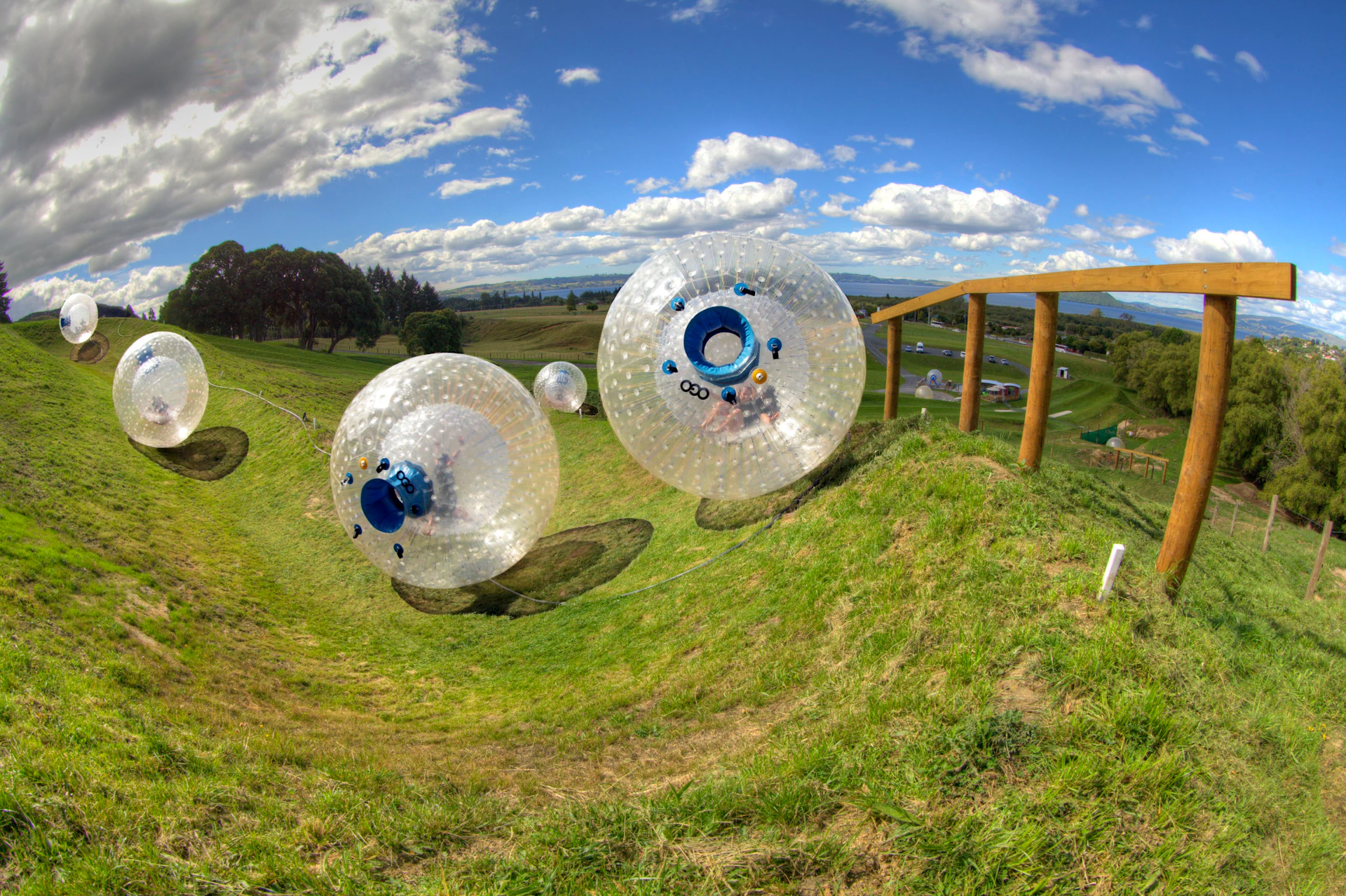 People are inside giant transparent zorb balls rolling down a grassy hill under a partly cloudy sky.