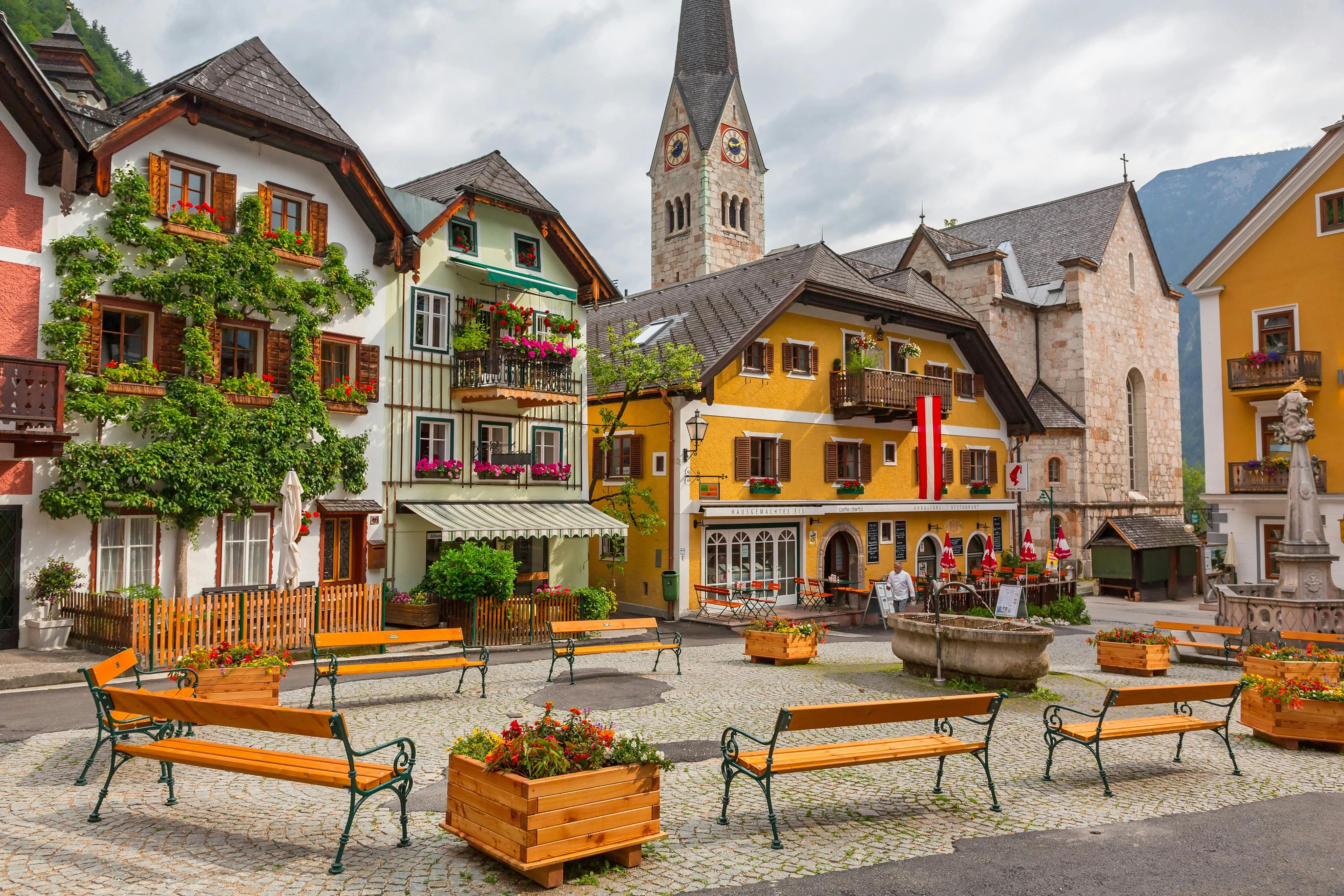 Ein malerischer Stadtplatz mit bunten Gebäuden, einem Kirchturm, Holzbänken, Blumenkübeln und Kopfsteinpflaster.