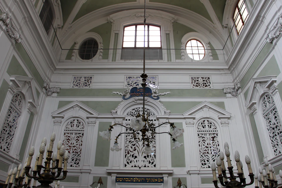 Ornate interior with arched windows, intricate carvings on white walls, and a central chandelier in a high-ceilinged room.