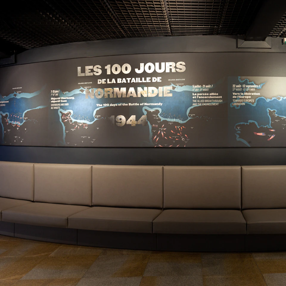 Display board reading "Les 100 Jours de la Bataille de Normandie" with a timeline and map. Curved gray seating in front.
