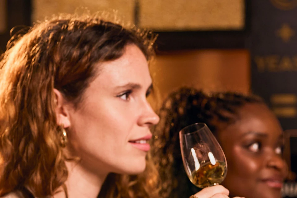 People seated at a table, sampling drinks from glasses in a warmly lit environment with a distillery decor.