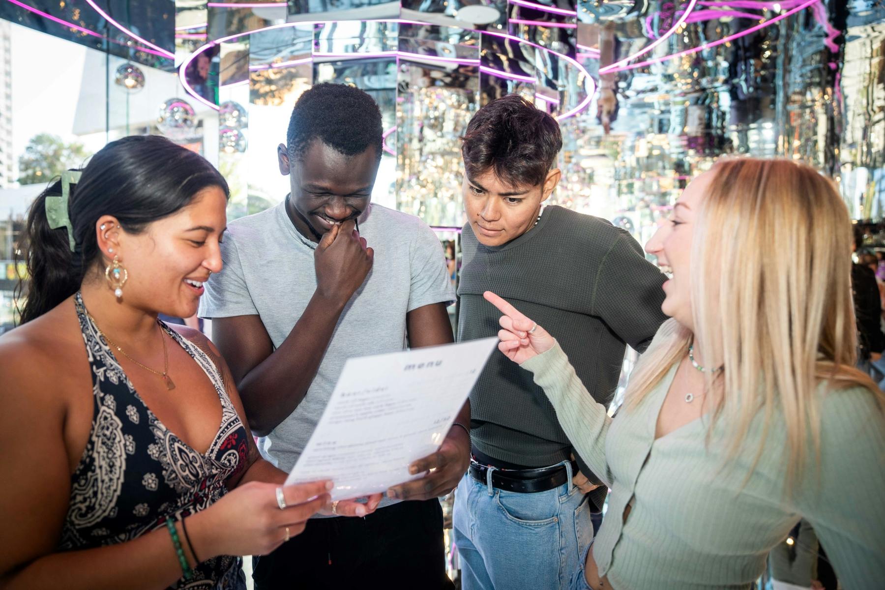 鏡のような壁と紫色のネオンライトが映し出された鮮やかな部屋で、4人がメニューを綿密に調べています。 一人がメニューを指差しています。