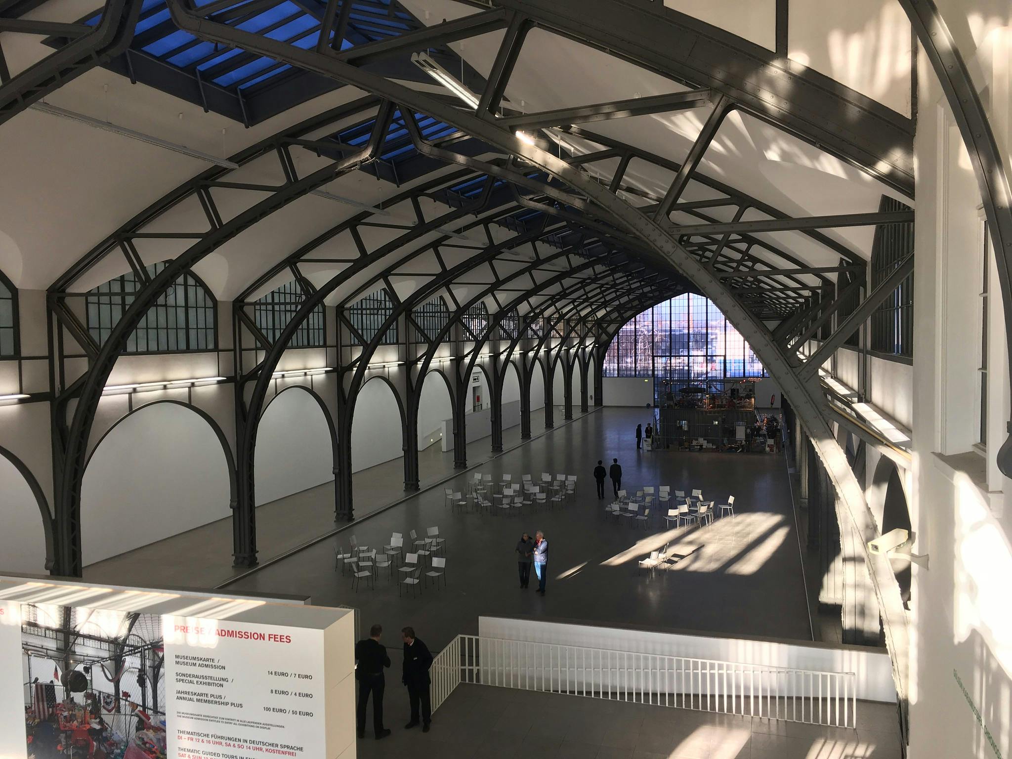 Prostorná hala v industriálním stylu s vysokými klenutými stropy, prázdnými židlemi a stoly a několika lidmi stojícími a chodícími kolem.