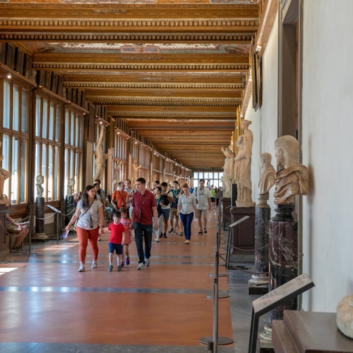 People walking in a corridor of a museum with statues on pedestals along the walls and large windows on the left side.