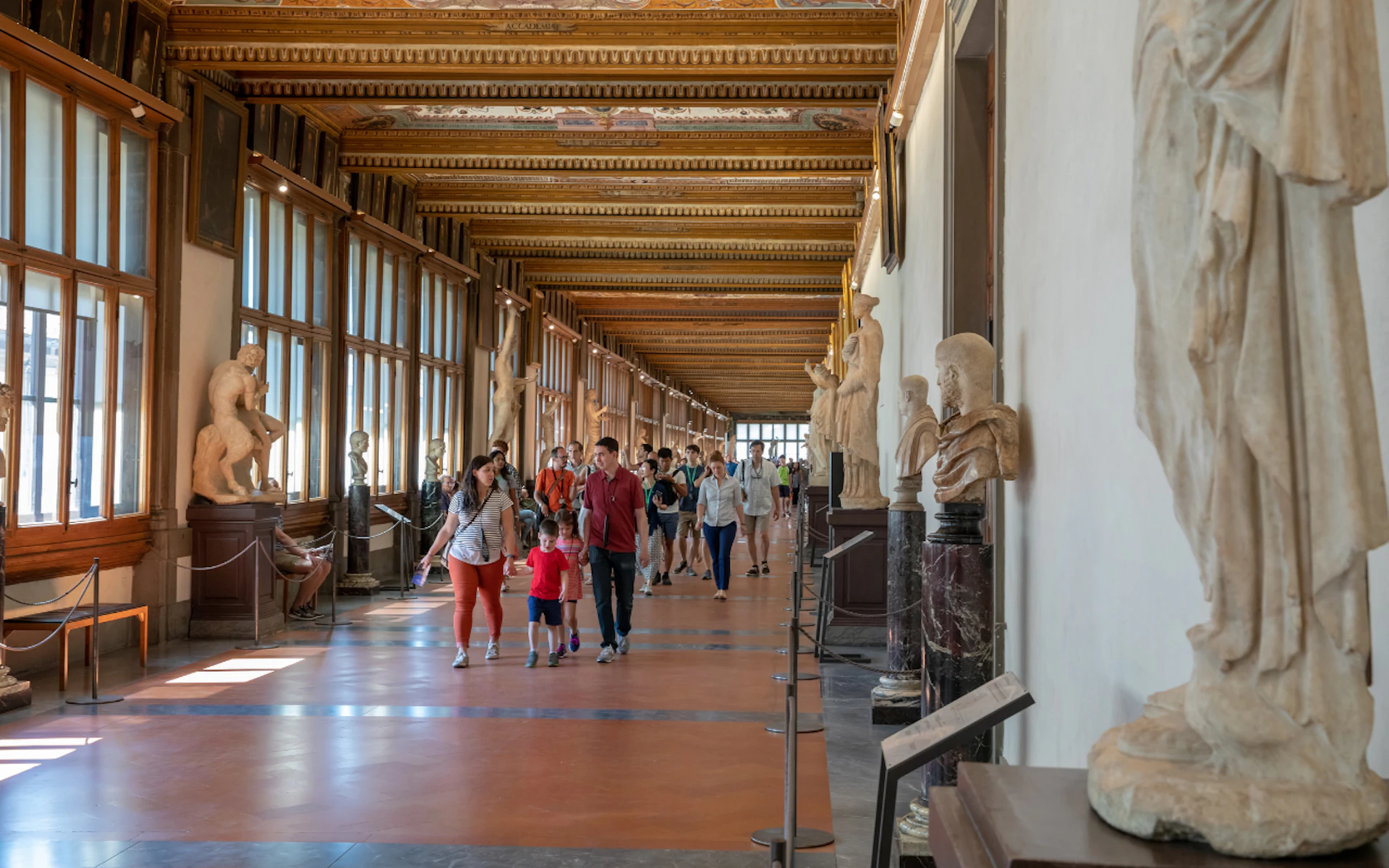 People walking in a corridor of a museum with statues on pedestals along the walls and large windows on the left side.