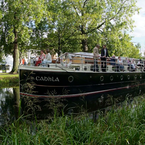 People are sitting and standing on a canal boat named "Cadhla," surrounded by green trees and grass reflecting in the water.