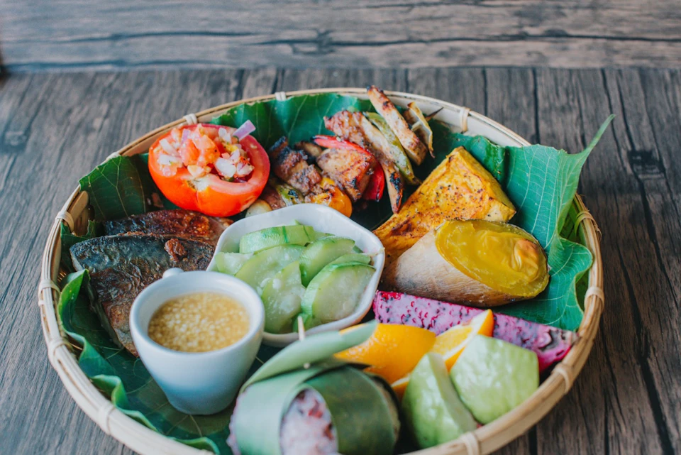 A basket lined with leaves containing various foods, including grilled meat, vegetables, fruits, and a dipping sauce.