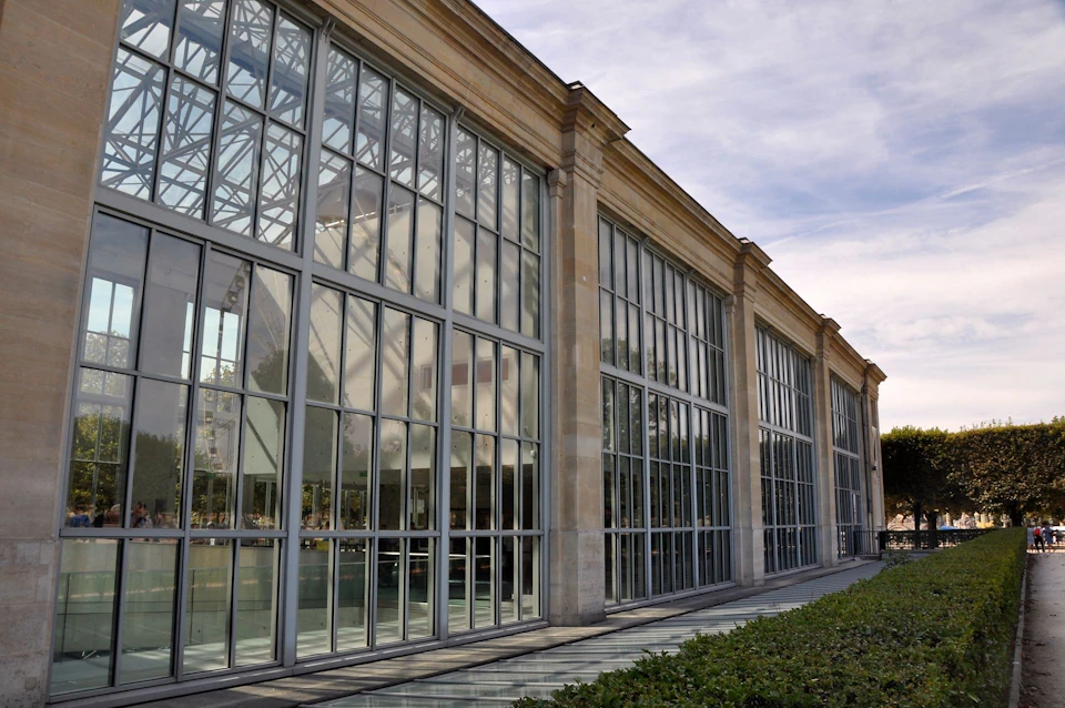 A glass-fronted building with a metal framework and large windows, under a cloudy sky, next to a neatly trimmed hedge.