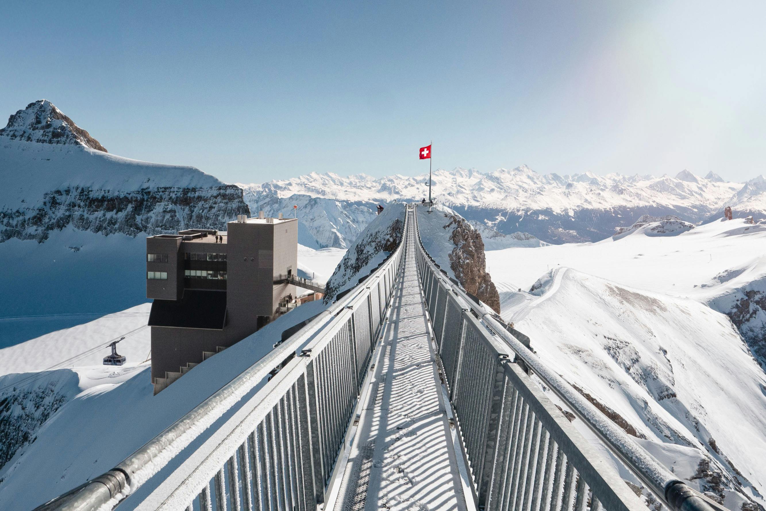 스위스 국기가 있는 봉우리로 이어지는 눈 덮인 보행자 다리는 산으로 둘러싸여 있으며 왼쪽에 건물이 있습니다.