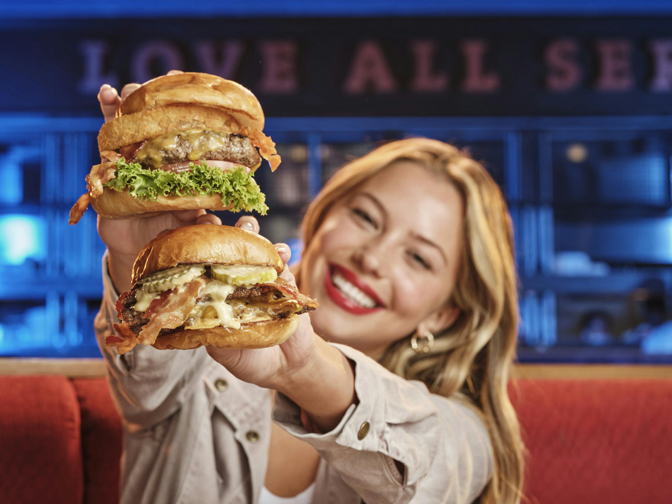 A woman with blonde hair and red lipstick smiles while holding up two cheeseburgers with lettuce and bacon.