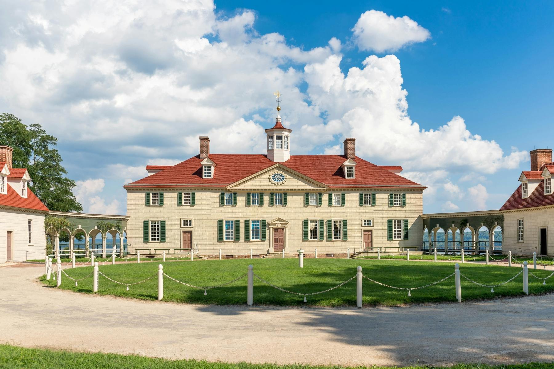 A historic mansion with a red roof, beige walls, and green shutters, set on a lawn with a blue sky and clouds above.
