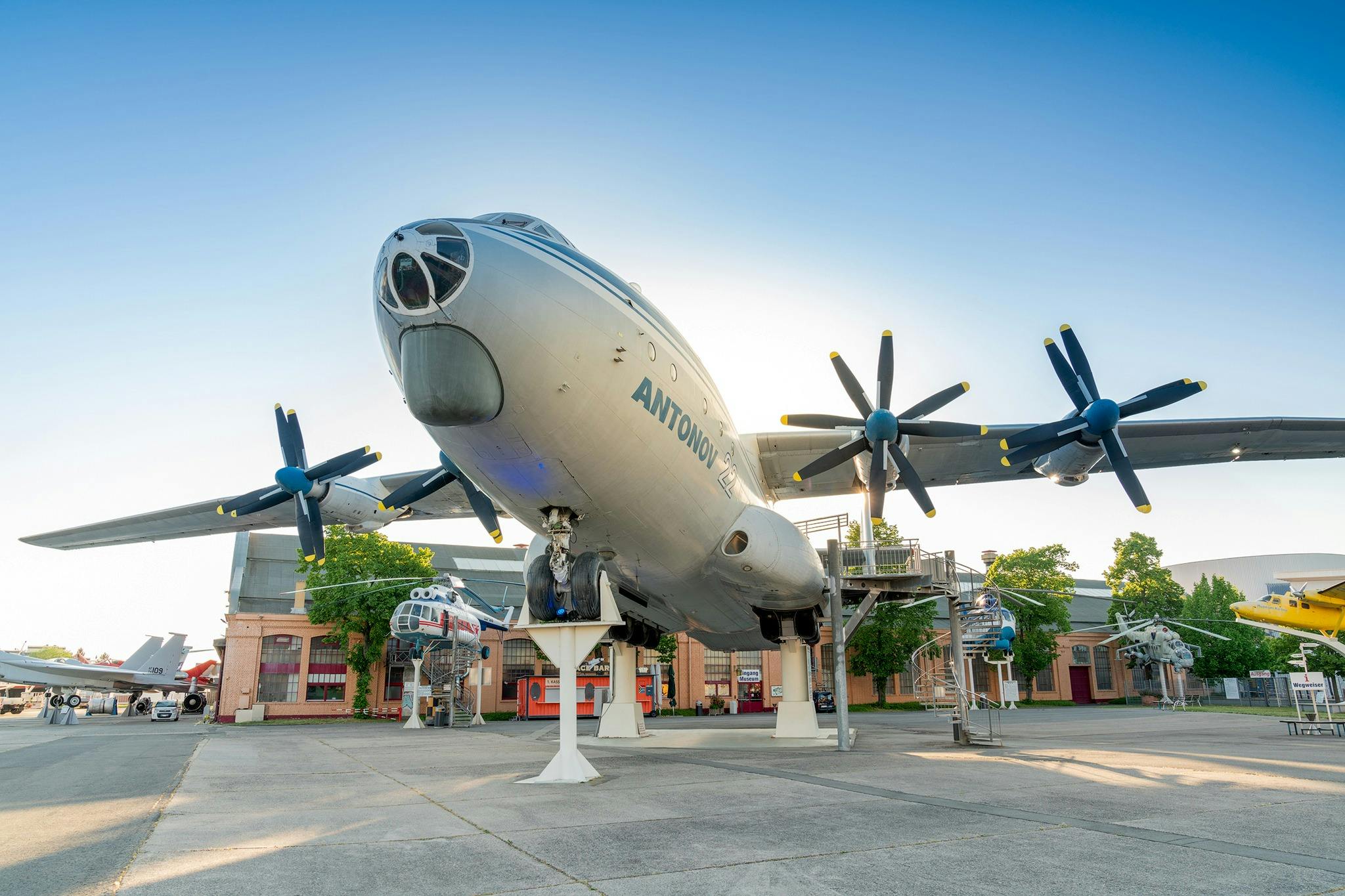 Un grand avion Antonov exposé en plein air sur des stands, entouré de plus petits avions et de bâtiments sous un ciel dégagé.