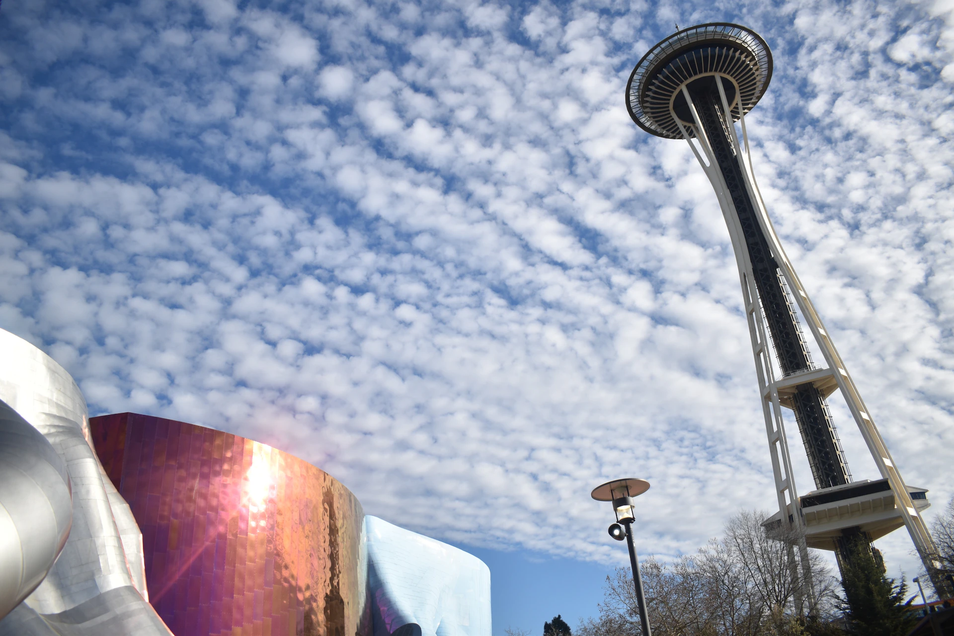 The image shows the Space Needle, a lamp post, and a reflective, curved building under a partly cloudy sky.