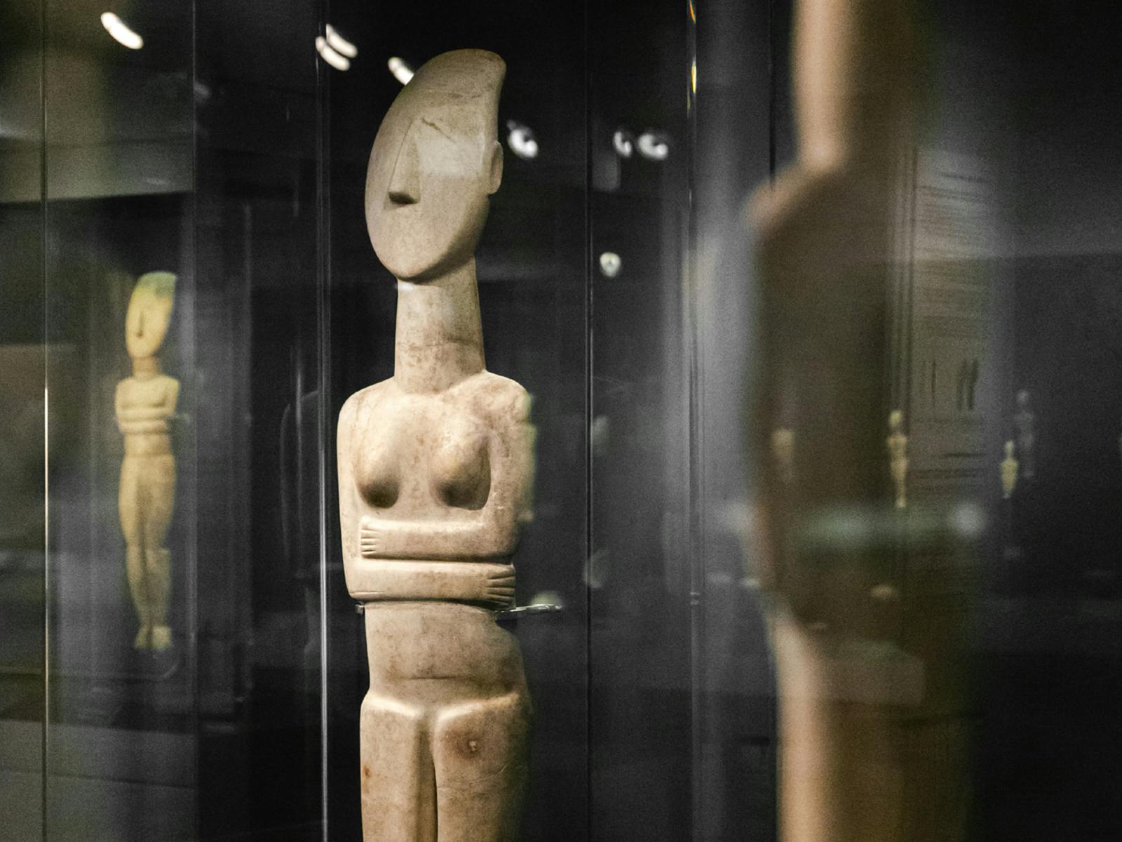 A stone statue with a stylized human form, arms crossed, displayed in a glass case within a museum.