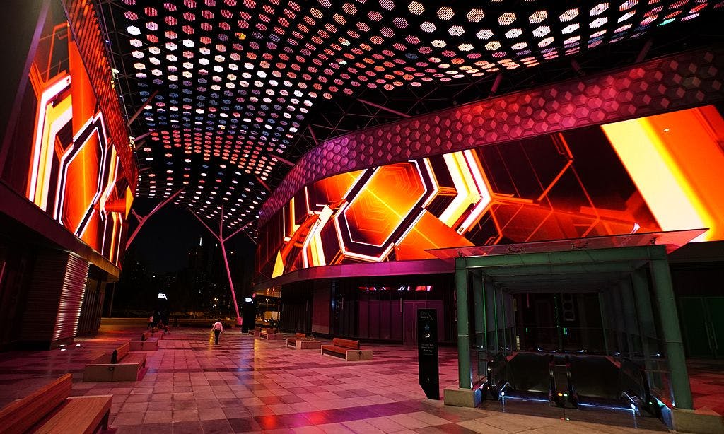 Udendørs plaza om natten, oplyst af levende geometriske lysdisplays på bygninger, med en tagbaldakin af sekskantede lys.