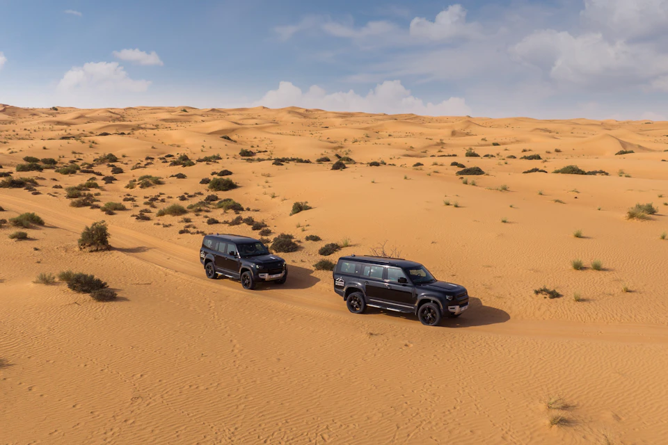 Two black SUVs drive through a vast, sandy desert with small bushes scattered across the landscape under a partly cloudy sky.