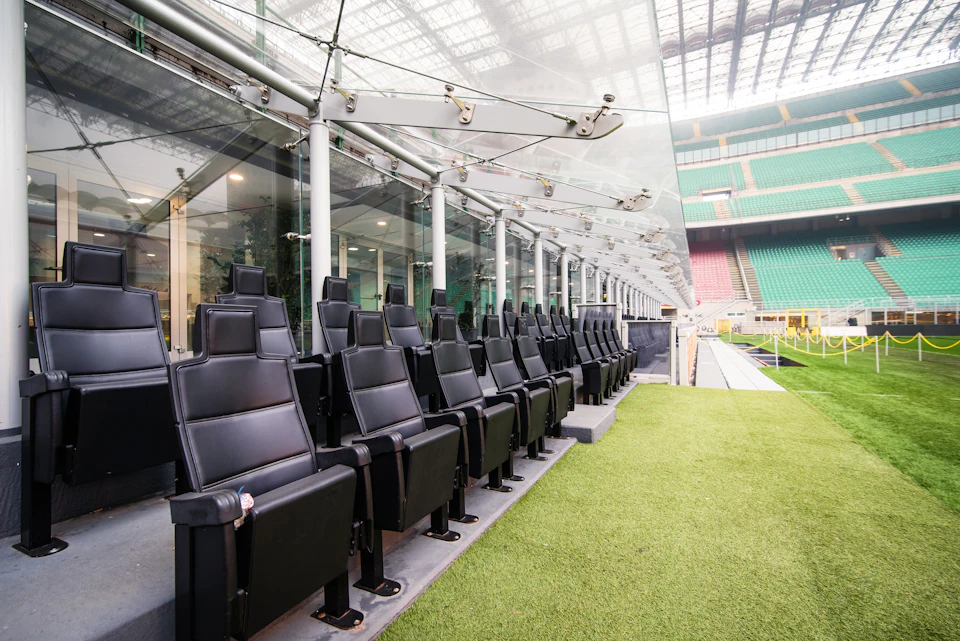Row of black seats in an empty, covered football dugout by a grassy field inside a large, multi-colored stadium.