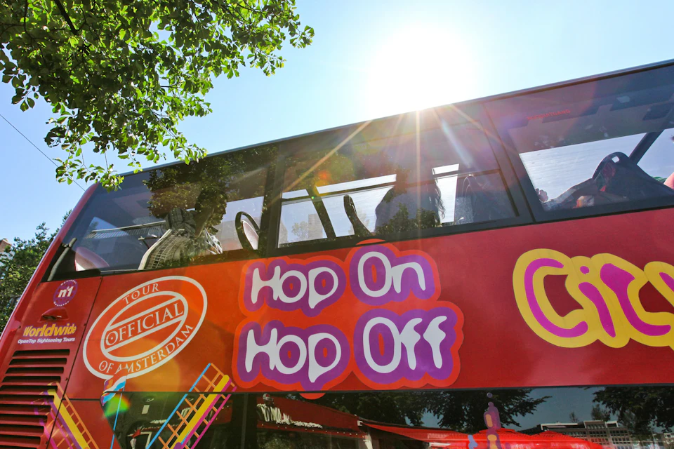 A double-decker tour bus with "Hop On Hop Off" signage, under a bright sun and partial tree canopy.