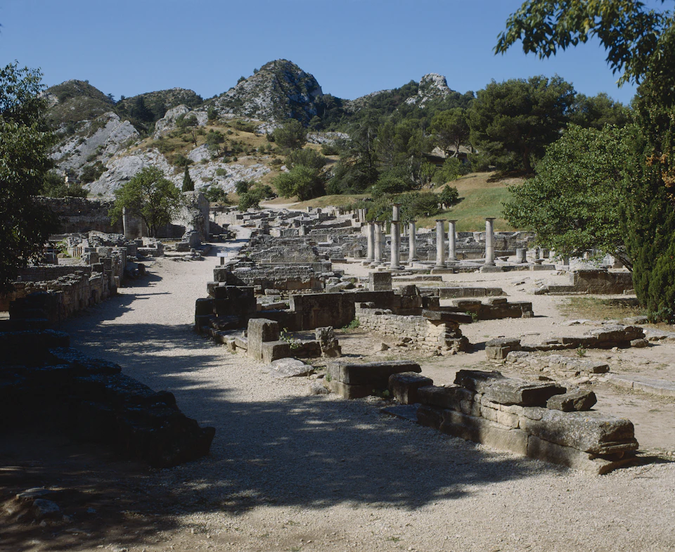 Ancient ruins featuring broken stone structures and standing columns, set against a backdrop of hilly, tree-covered terrain.