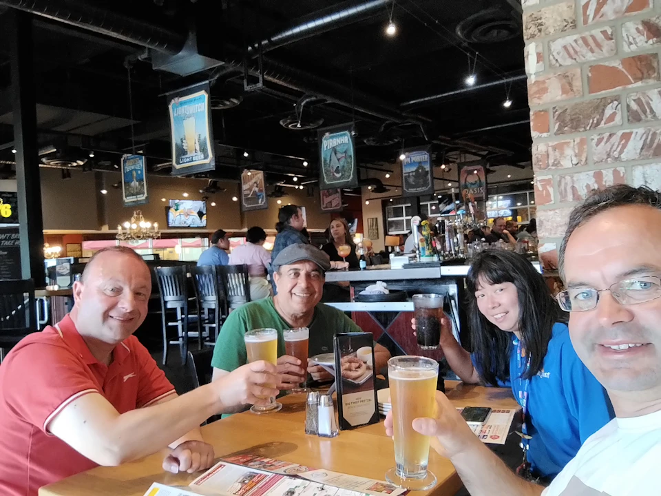 Four people sit at a restaurant table, smiling and holding drinks. The background shows other patrons and bar area.