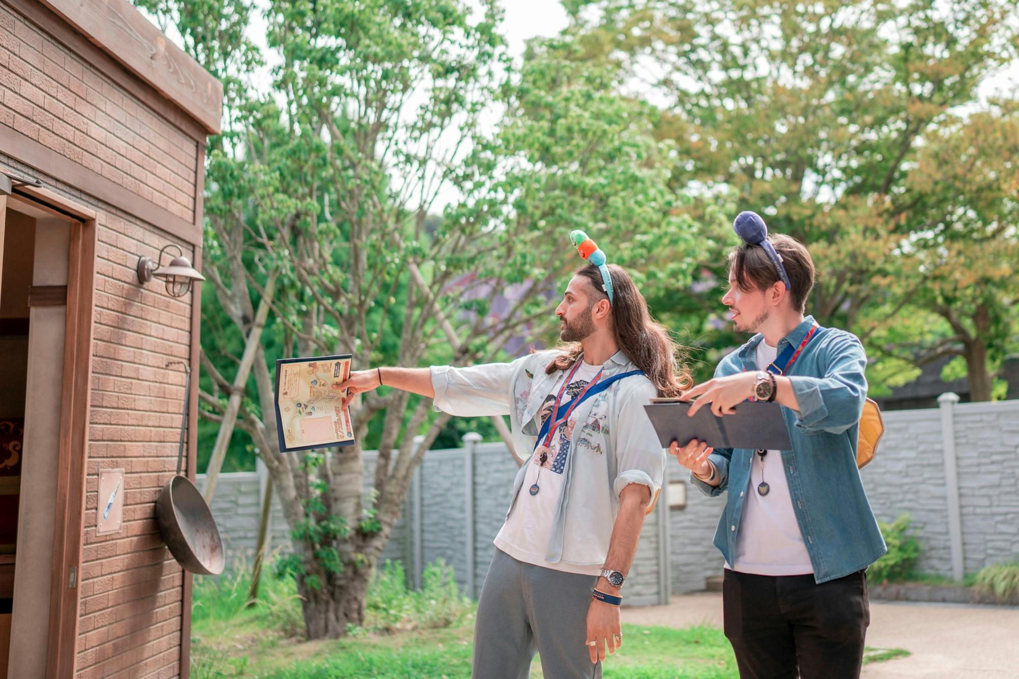 Two people outdoors holding a map and tablet, with headbands featuring small decorations, standing near trees and a wall.