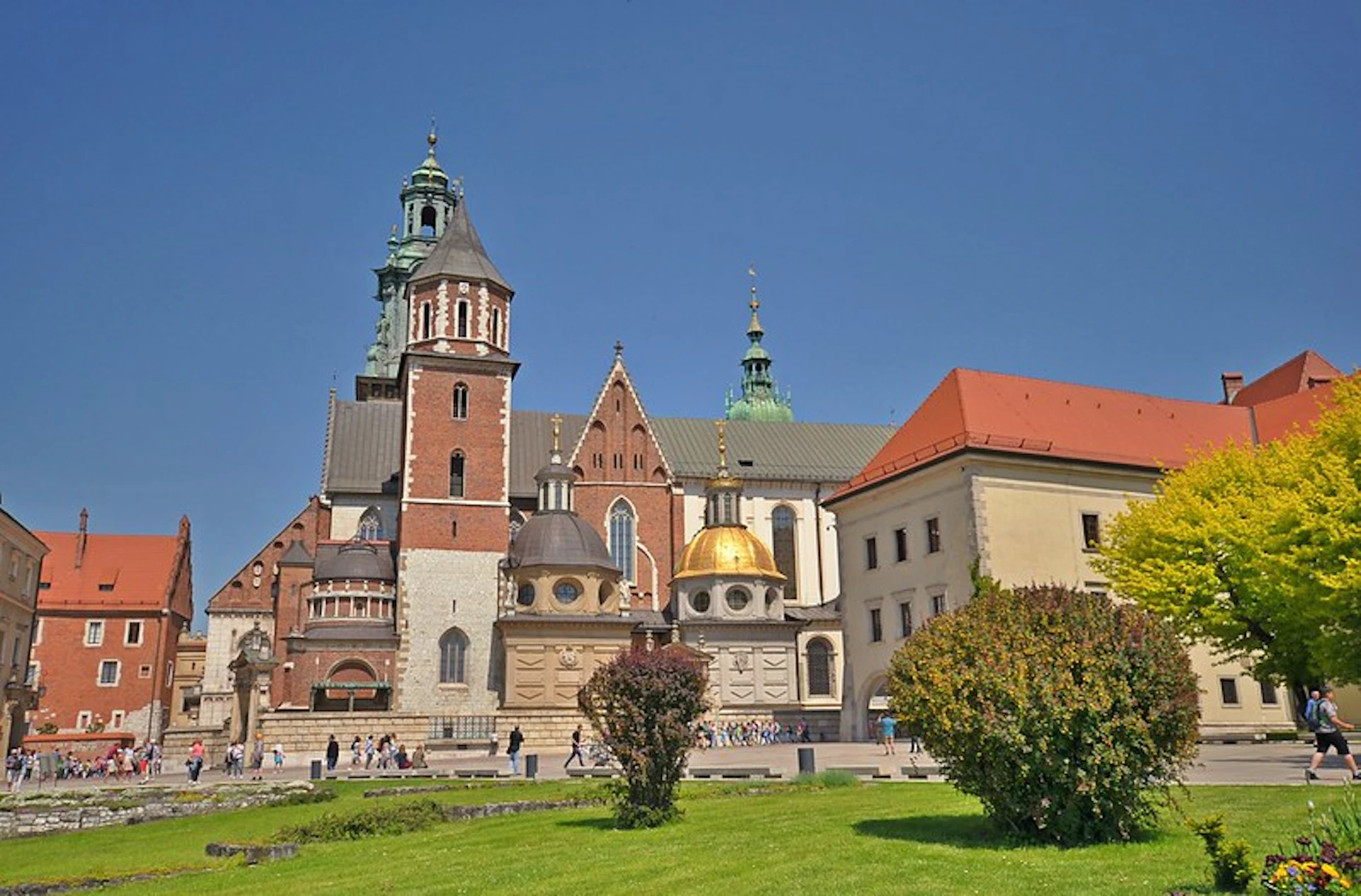 A historical building with multiple towers and domes, surrounded by a grassy area with bushes, against a clear blue sky.