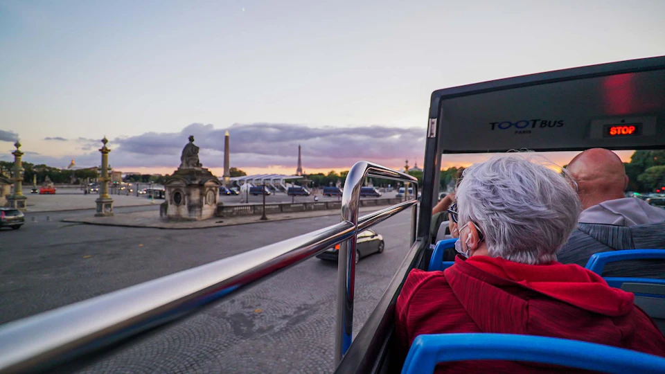 A person in a red jacket rides an open-top bus, overlooking historical monuments and a cloudy sunset sky.
