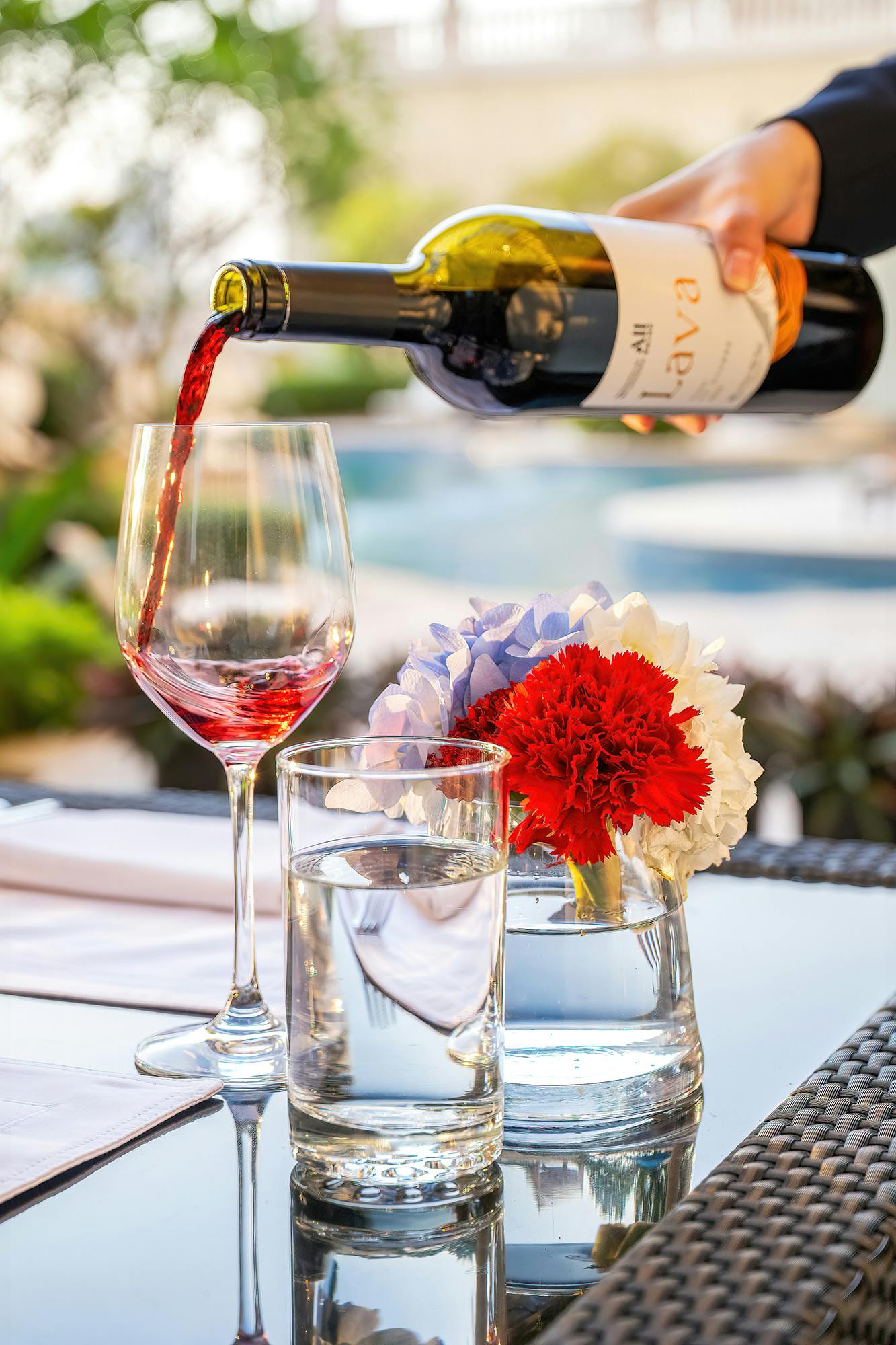 A glass of red wine being poured beside a glass of water and a vase with red and white flowers on a table outdoors.