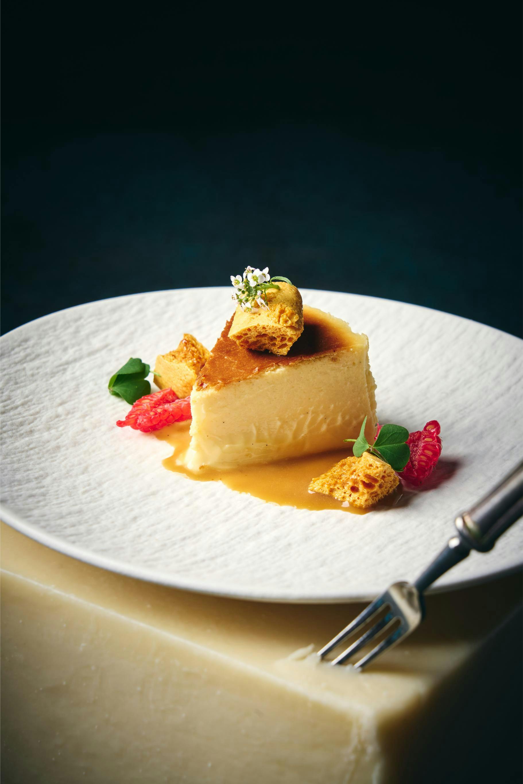 A dessert plated with custard, sauce, honeycomb, raspberries, and garnish on a white textured plate with a fork nearby.