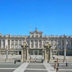 Stor utsmyckad byggnad med kolumner och detaljerade fasader bakom en inhägnad innergård. Människor går i förgrunden under en klar himmel.
