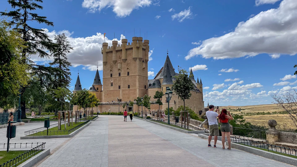 A grand castle with tall towers and spires under a blue sky with scattered clouds, tourists in the foreground taking photos.