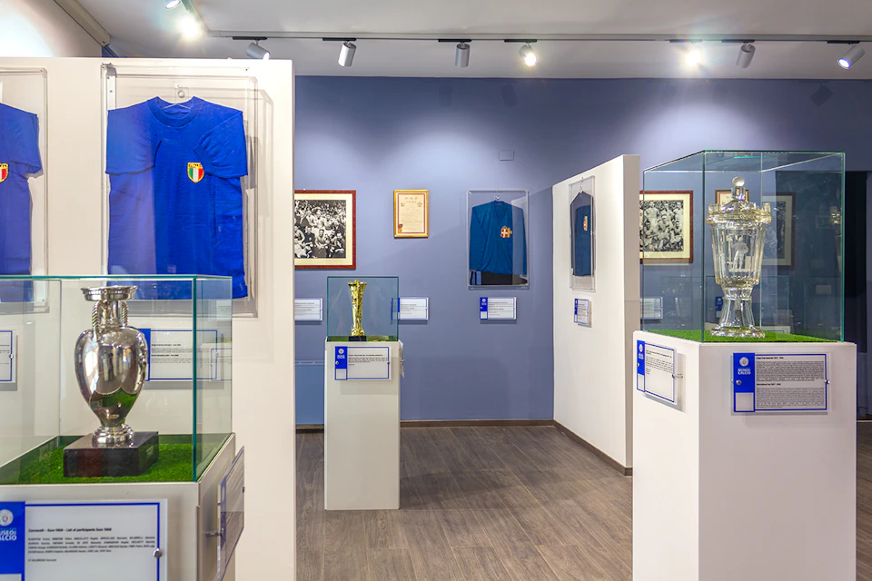 Museum exhibit displaying sports memorabilia, including blue jerseys, framed photographs, and a gold trophy in glass cases.