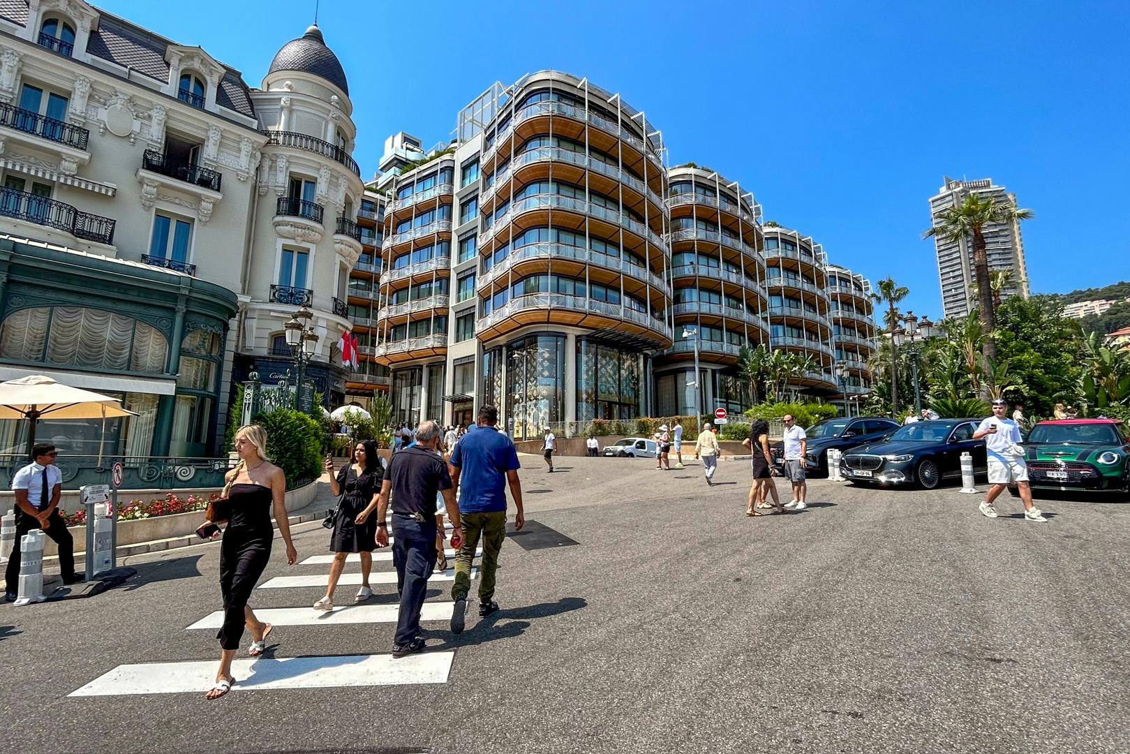 People walking on a street near modern and historic buildings under a clear blue sky.