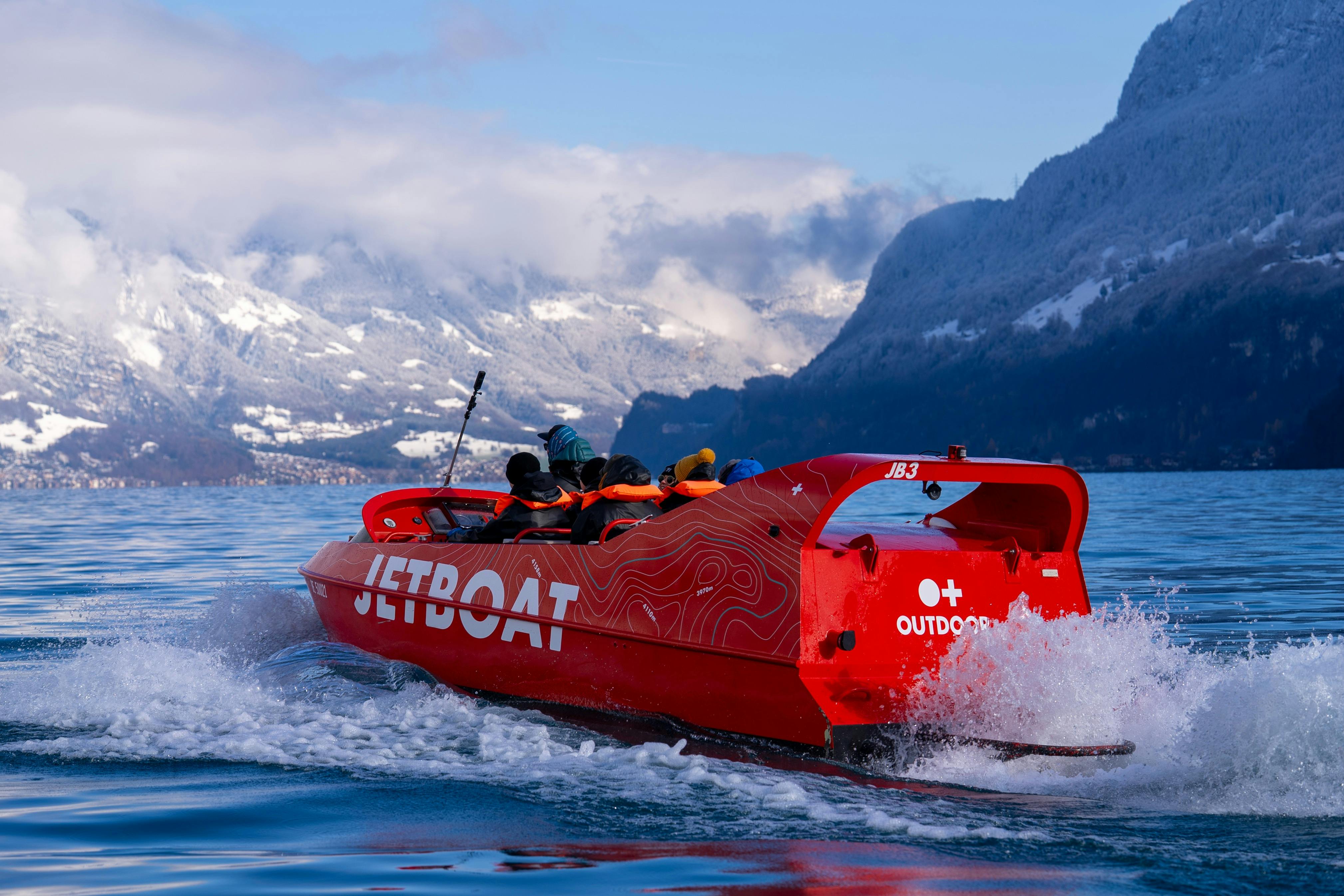 A red jet boat with people onboard speeding across a lake, surrounded by snow-covered mountains under a partially cloudy sky.