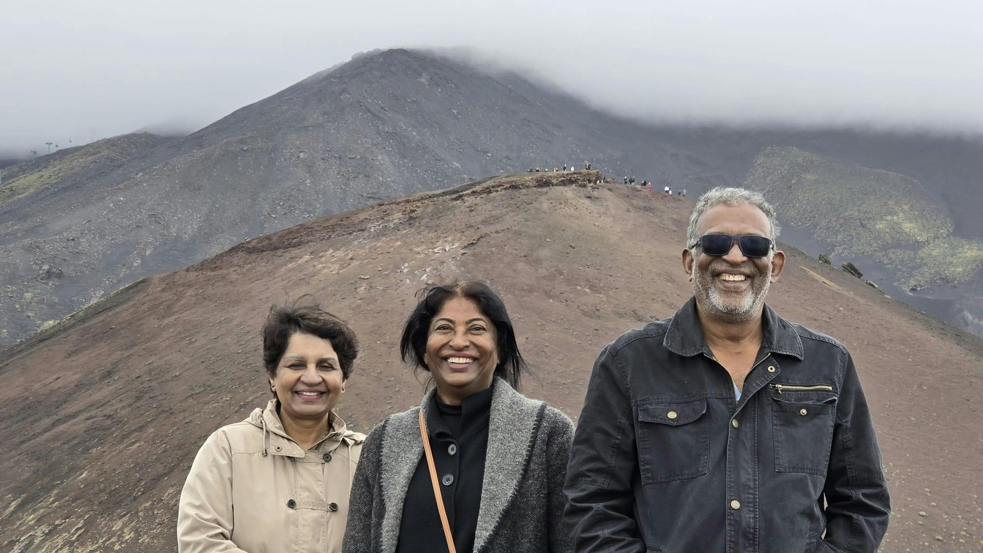 Τρεις άνθρωποι χαμογελούν μπροστά σε ένα βραχώδες, ορεινό τοπίο με ομίχλη, κάτω από έναν συννεφιασμένο ουρανό.