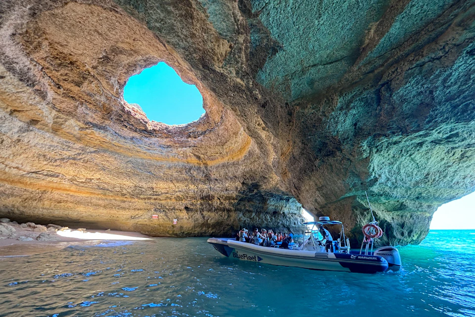 A boat with people inside a cave with a large circular opening at the top, revealing the sky, and a small beach visible.