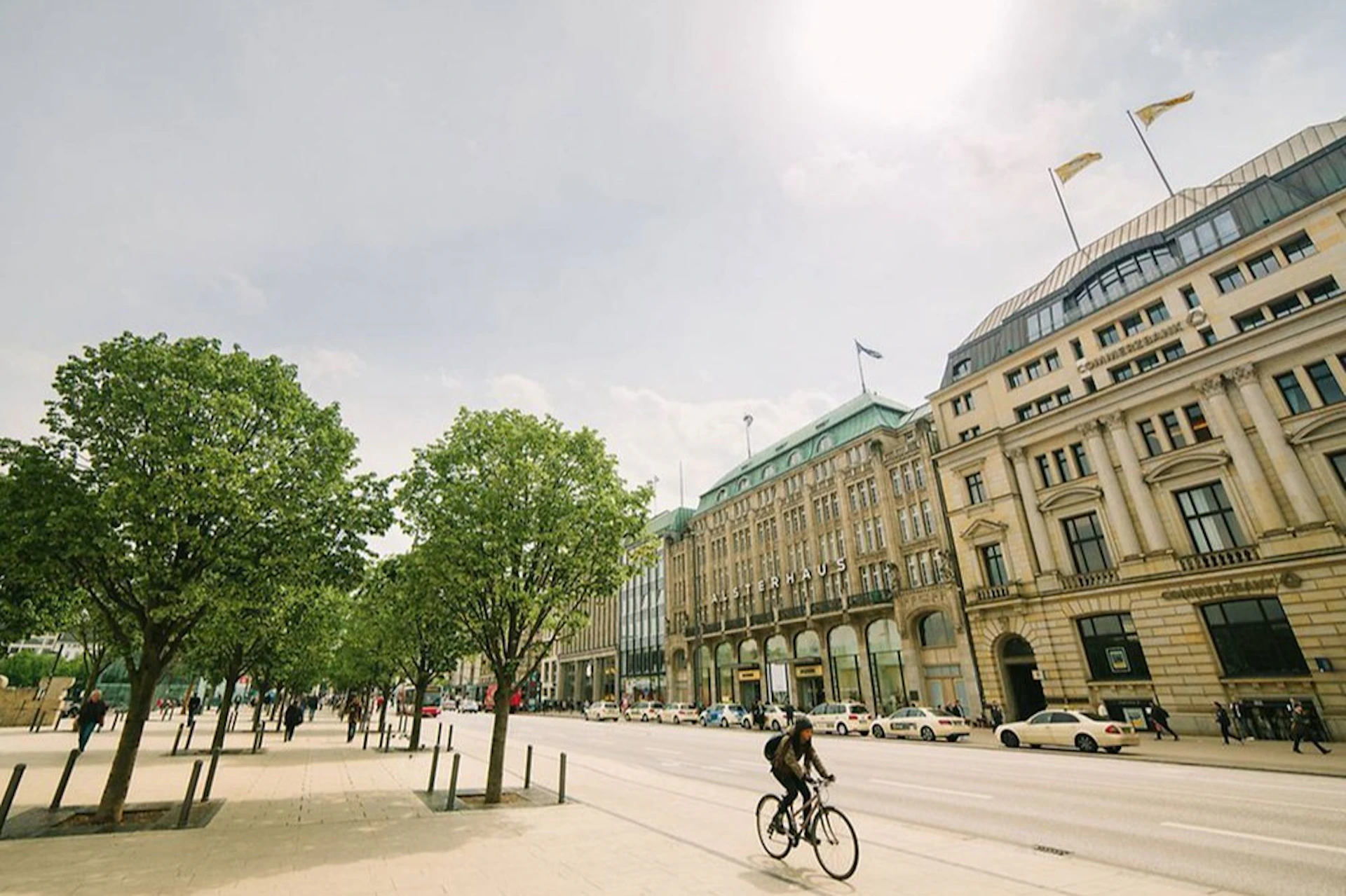 A cyclist rides along a wide street lined with trees and historic buildings under a bright, partly cloudy sky.