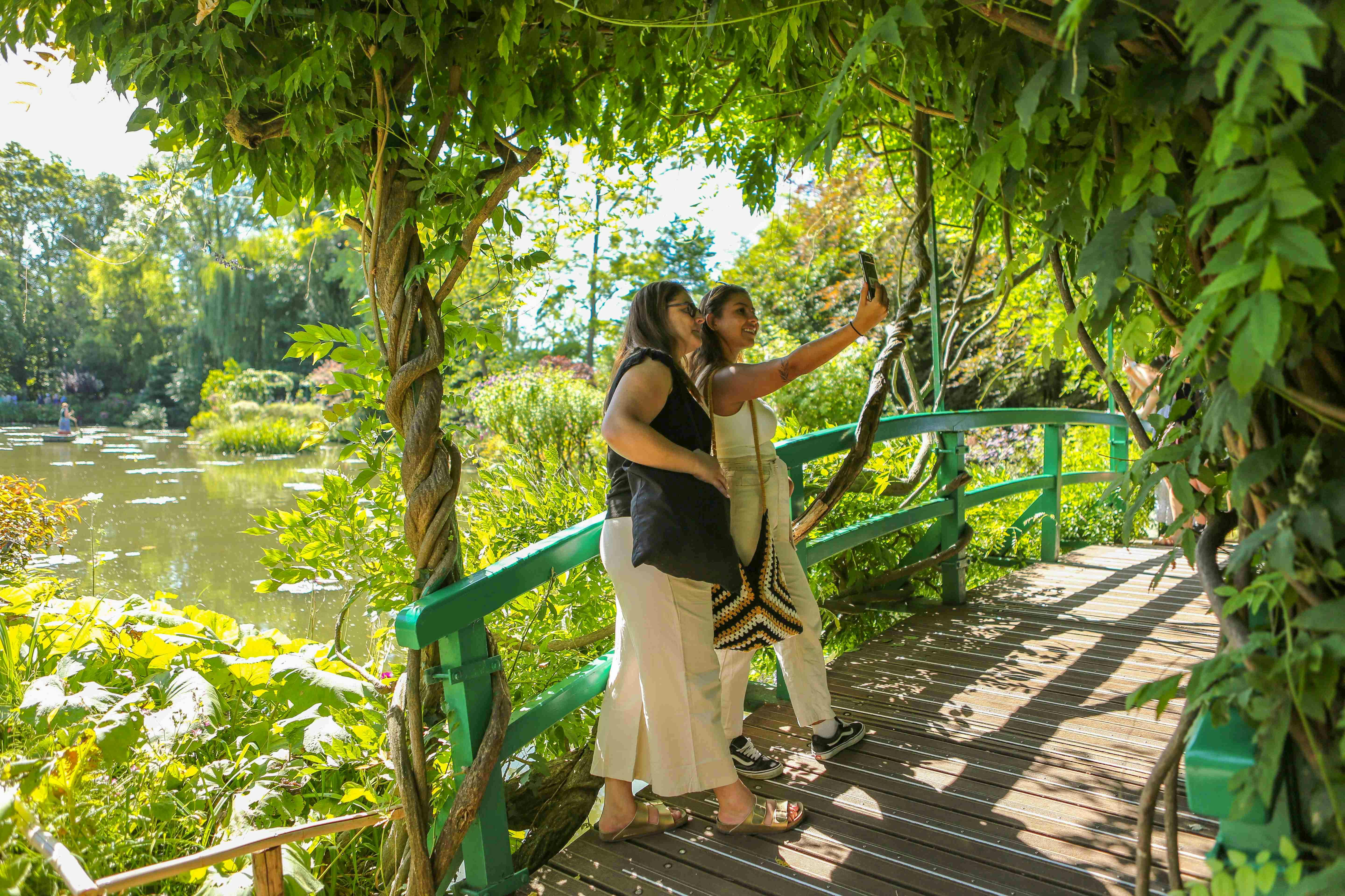Δύο γυναίκες στέκονται σε μια ξύλινη γέφυρα που περιβάλλεται από πλούσια βλάστηση, βγάζοντας μια selfie κάτω από μια αψίδα καλυμμένη με αμπέλια.