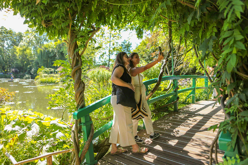 Two women stand on a wooden bridge surrounded by lush greenery, taking a selfie under a vine-covered arch.