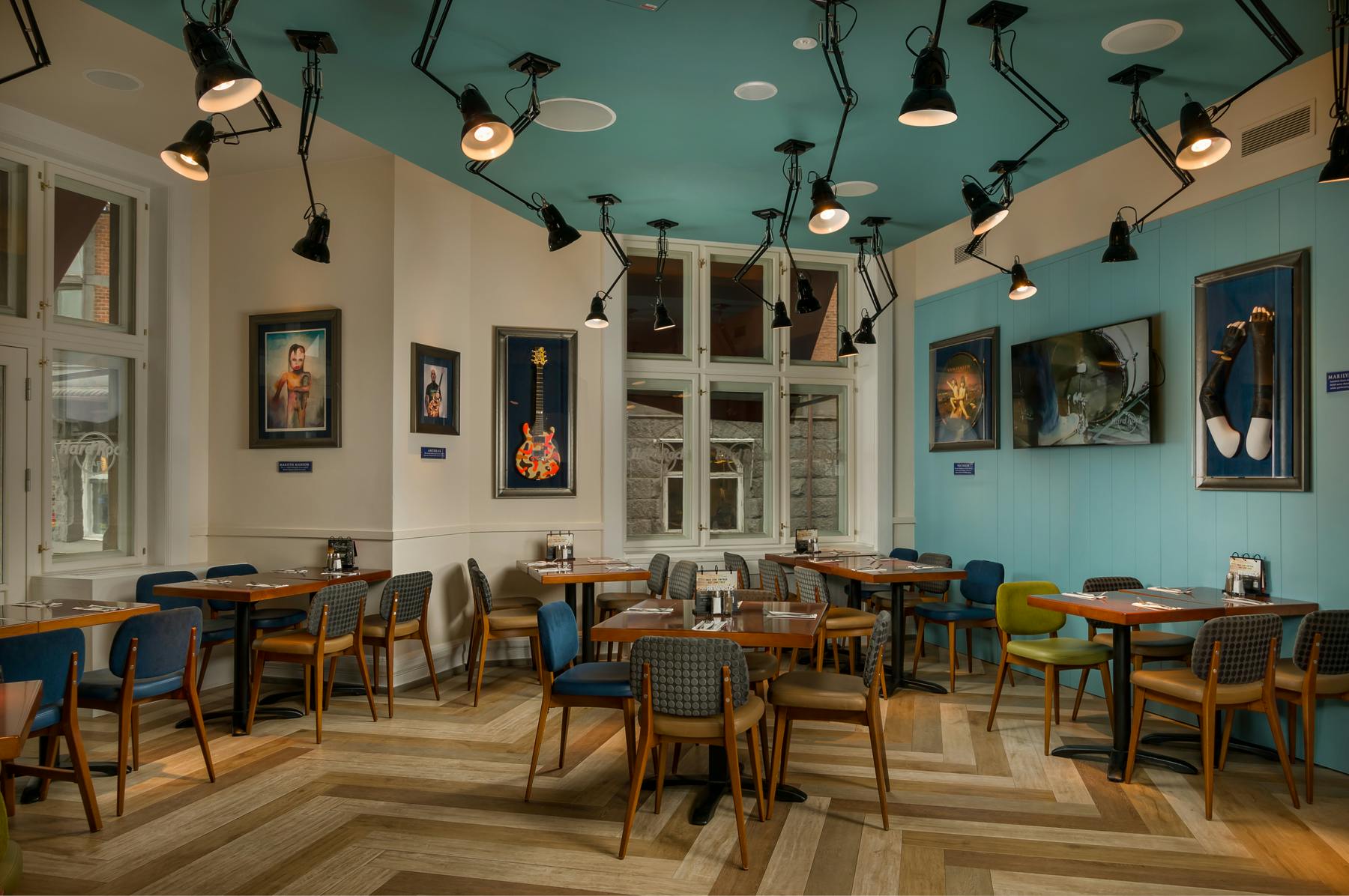 현대적인 인테리어로 꾸며진 세련되고 빈 카페에는 나무 테이블, 다양한 색상의 의자, 벽면 예술, 수많은 천장 조명이 있습니다.