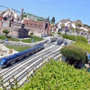 Persone che osservano un grande plastico ferroviario all'aperto con edifici e paesaggi dettagliati sotto un cielo azzurro e limpido.