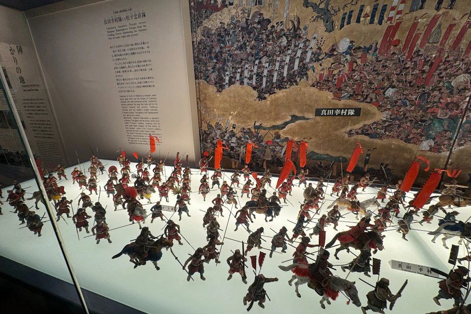 A display of miniature soldiers in battle formation with red flags in front of historical battle artwork and text in Japanese.