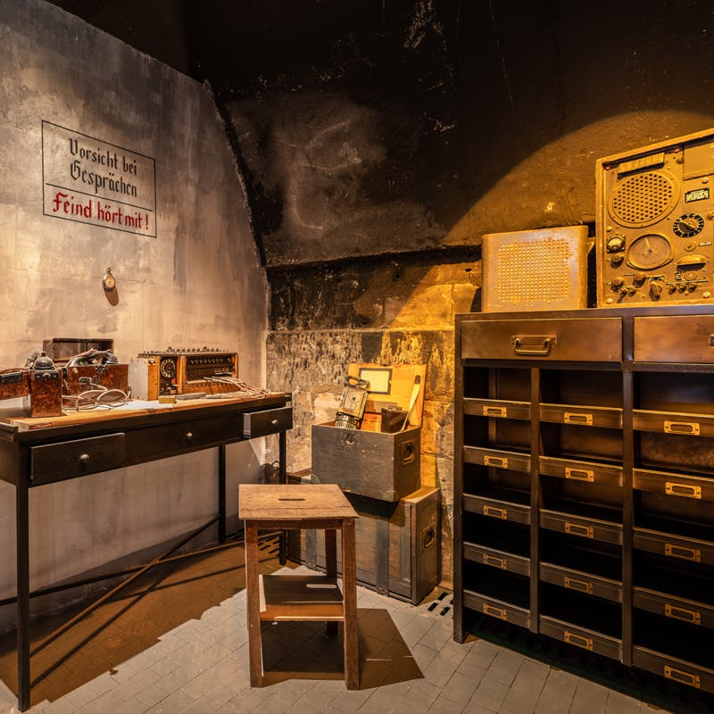 Stanza poco illuminata con apparecchi radiofonici d'epoca su una scrivania, uno sgabello, scatole e un armadietto con cassetti etichettati.