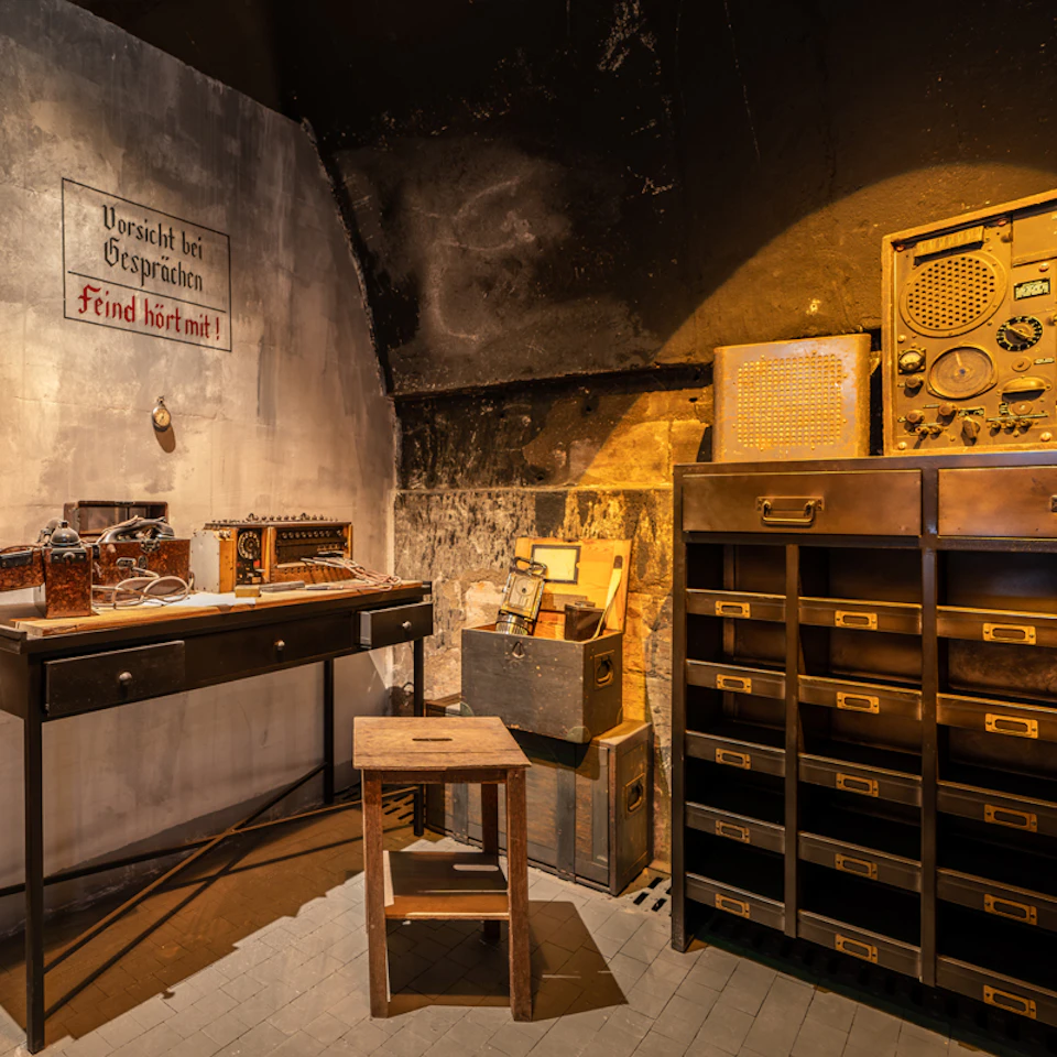 Dimly lit room with vintage radio equipment on a desk, a stool, storage boxes, and a cabinet with labeled drawers.