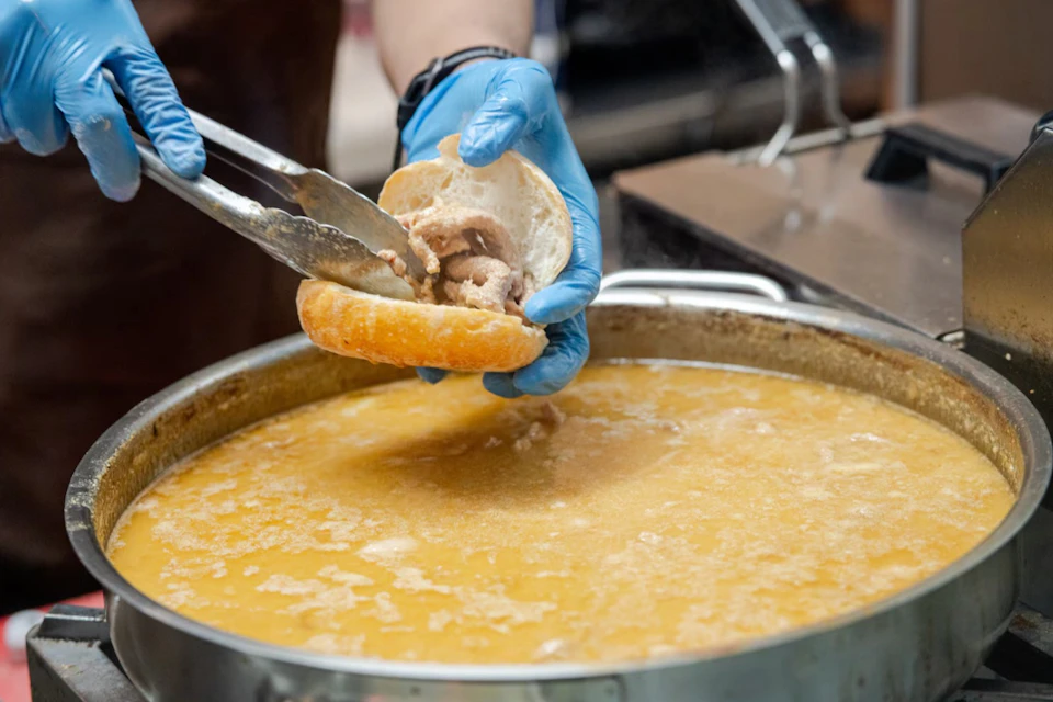 A person wearing blue gloves uses tongs to place meat into a sandwich over a large pot of broth.