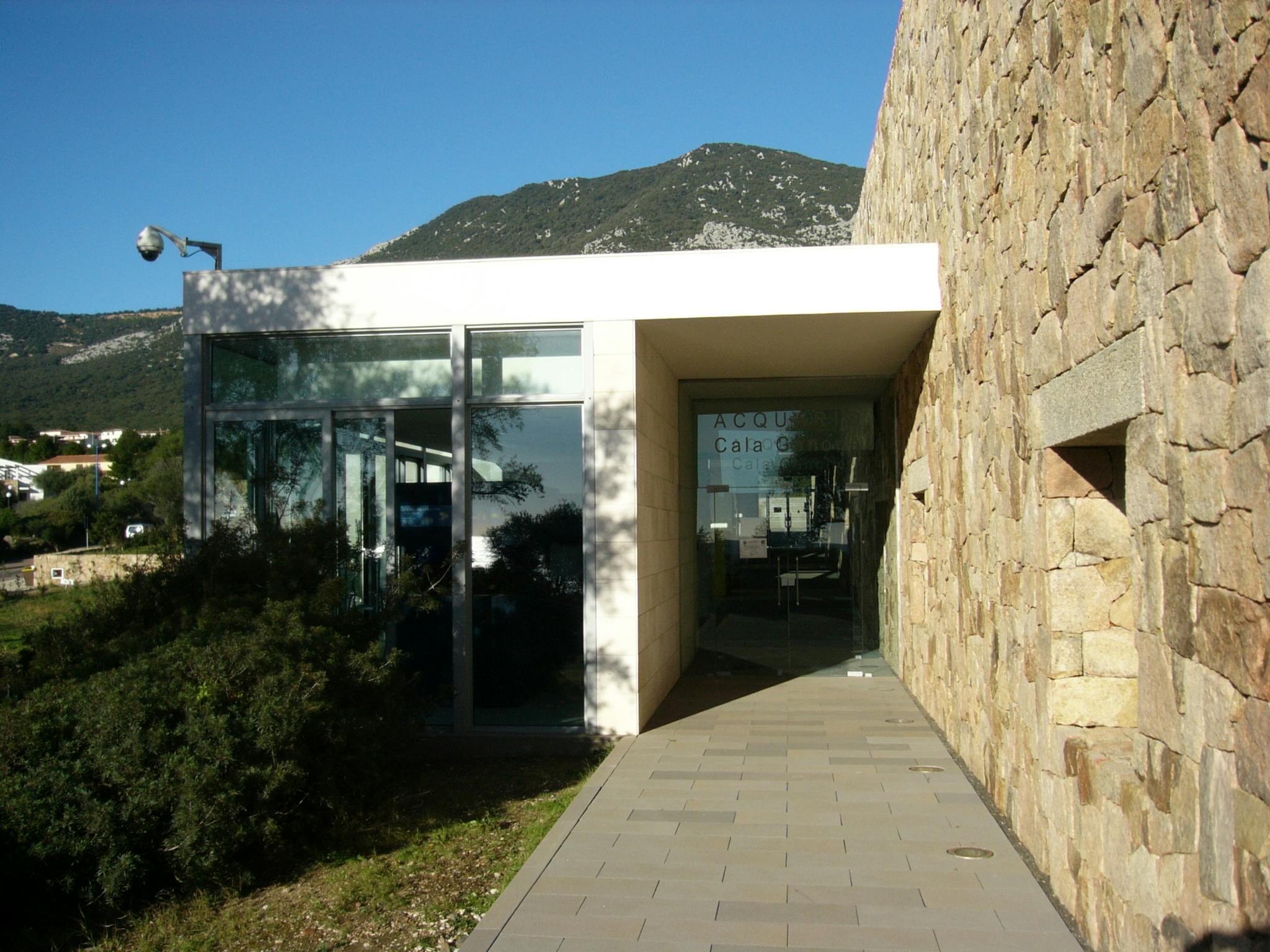 Moderne bygning med glasvægge, stenudvendig og en brolagt sti, der fører til en glasindgangsdør. Bjerg i baggrunden.